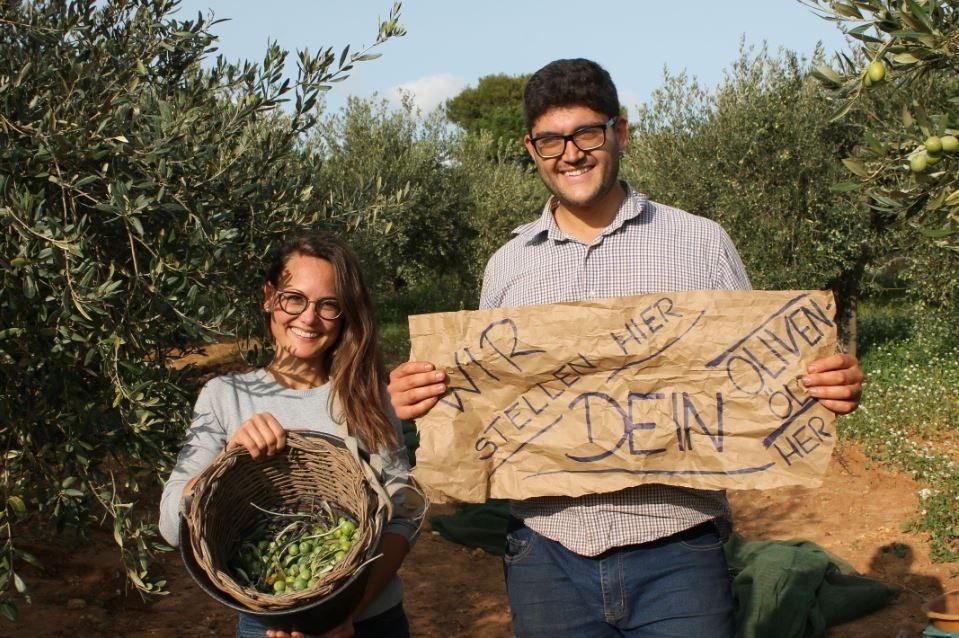 Zwei junge Erzeuger im Olivenhain halten ein Schild „Wir stellen hier dein Olivenöl her